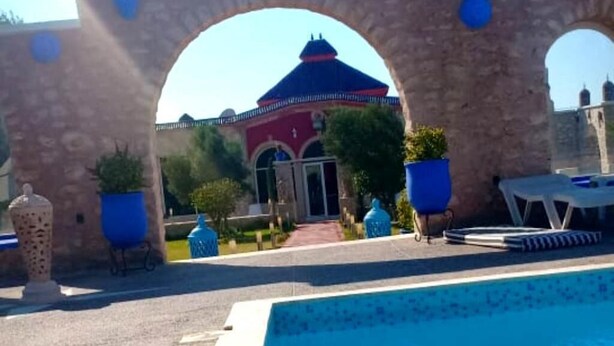 An outdoor swimming pool area with a stone archway, potted plants, and a building with a dome roof in the background. - Charming villa with pool 4160 (Essaouira)