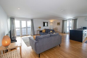 A spacious living area with a grey sofa, wooden flooring, a small table with a lamp, and a kitchen area with a black countertop.