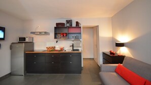 Un espace de vie compact comprenant un coin cuisine, un canapé avec un coussin rouge et une lampe fixée au mur.