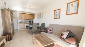 Un salon moderne avec un canapé, une table basse et des fauteuils. On aperçoit, au fond, une cuisine avec des meubles en bois et un coin repas.