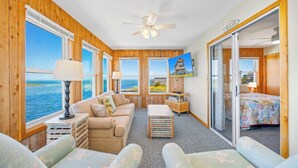 A room with a wooden interior, a ceiling fan, a bed with a colorful bedspread, a television, and a view of the sea through large windows. - Soundfront Home w/ Private Boat Dock, Game Room, Windsports Access, & More! (Avon)