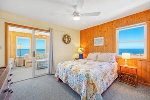 Um quarto com cama, mesa de cabeceira, janela com vista para o oceano, ventilador de teto e porta que dá para uma varanda.