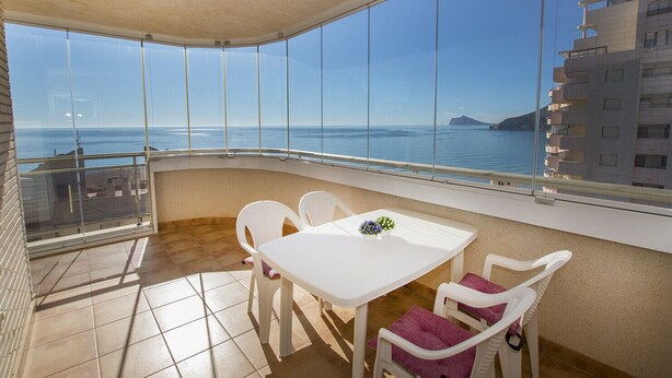 A balcony with a white table and chairs, a small flower arrangement, and a view of the sea and buildings.