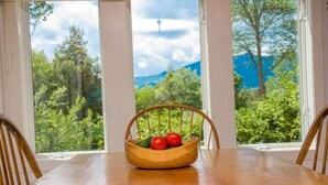 Ein Esstisch mit einem Holzstuhl, ein Korbkistchen mit Holzobst und Tomaten sowie ein Blick auf Grünflächen und Berge durch große Fenster.
