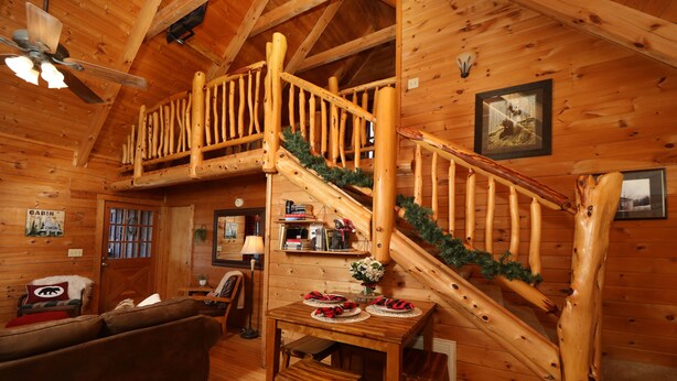 Interior de una cabaña con una escalera de madera, una mesa de comedor con un adorno navideño, un sofá y un ventilador de techo.