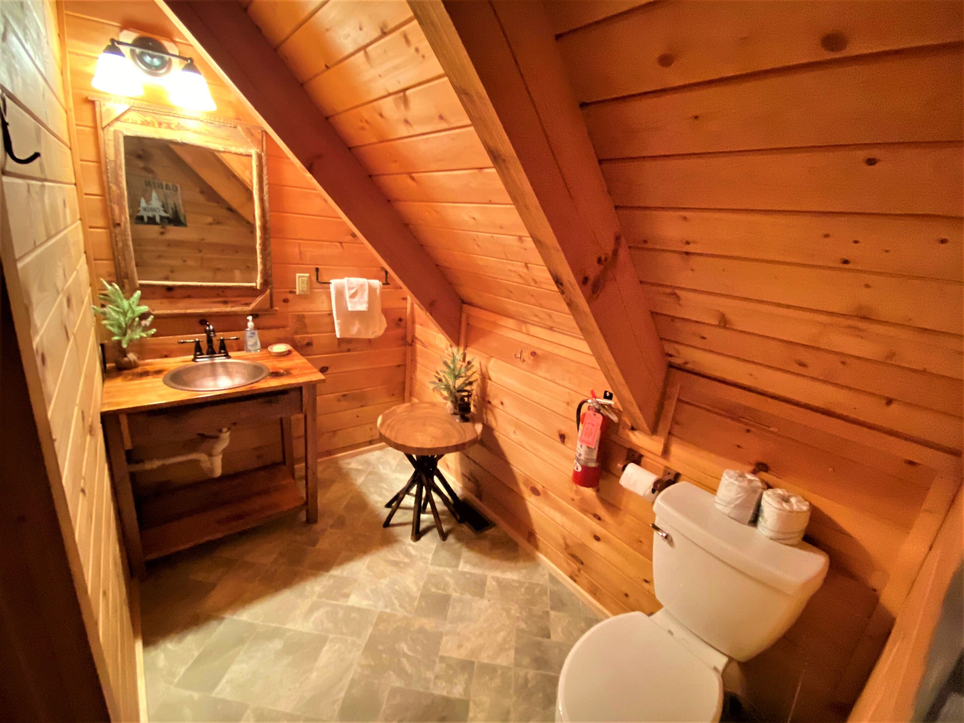 Baño de cabaña de madera con lavamanos, espejo, inodoro y una pequeña mesa redonda.