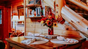 Un interior de madera con una mesa de comedor preparada para dos, una estantería llena de libros y una escalera de madera.
