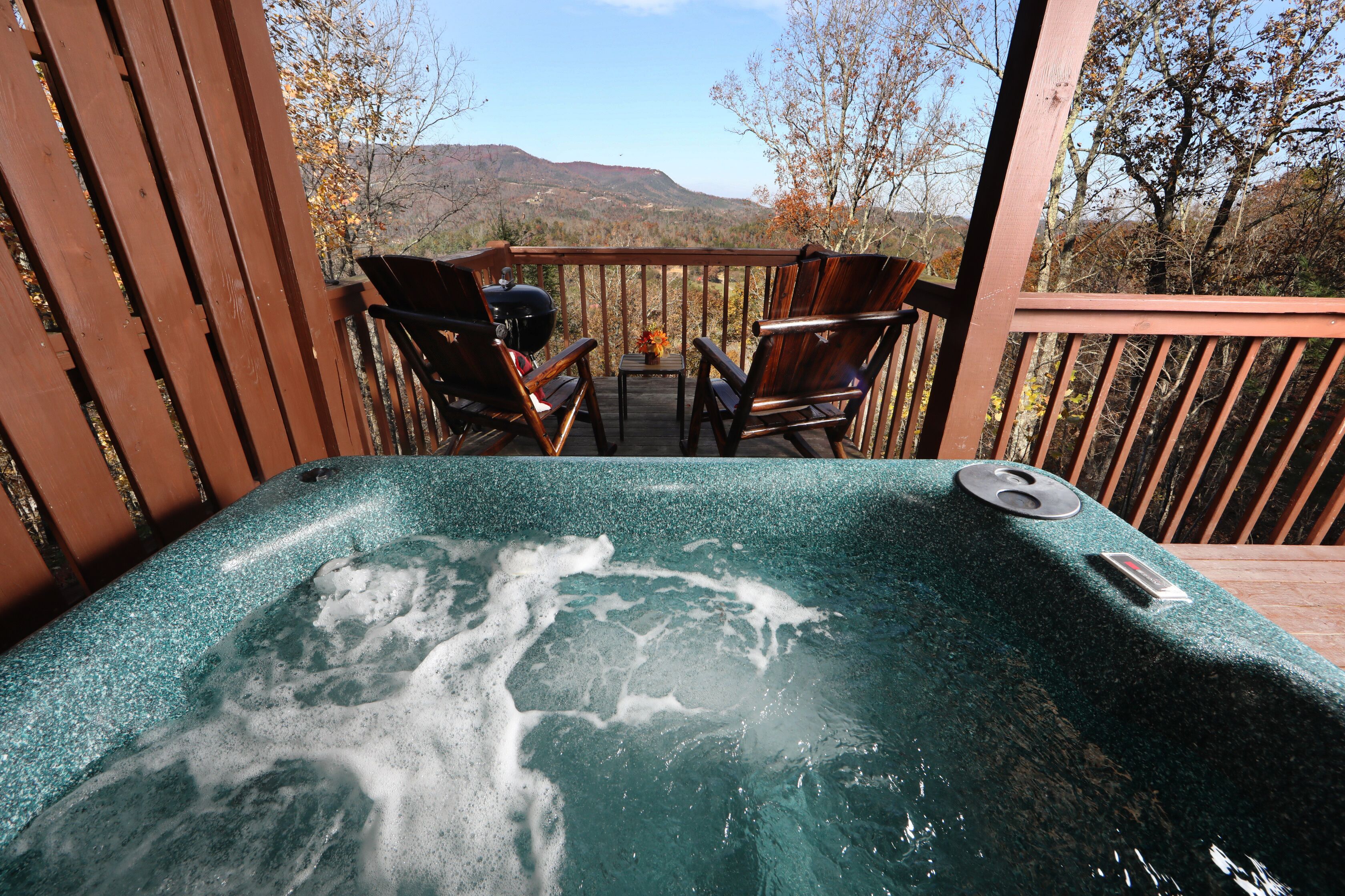 A hot tub on a wooden deck with chairs and a table, overlooking a mountainous landscape.