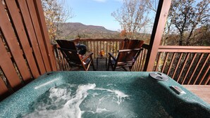 Un jacuzzi en una terraza de madera con sillas y una mesa, con vista a un paisaje montañoso.