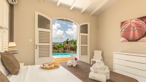 Ein Schlafzimmer mit Bett, Nachttisch mit Frühstücksgebäck und -getränken, Handtuchhalter mit Handtüchern sowie Blick auf Pool und Grünflächen durch offene Fensterläden.