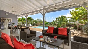 Ein überdachter Außenbereich mit Korbmöbeln, einem Glastisch und Blick auf Pool und Garten.