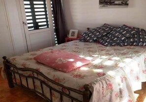 A bedroom with a bed, floral bedding, a pink pillow, patterned pillows, a small bedside table with a picture frame, and a window with blinds.