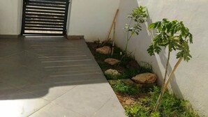 Ein gepflasterter Weg führt zu einem schwarzen Tor, ein kleiner Garten mit Steinen und einigen Pflanzen sowie eine weiße Mauer.