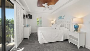 A bedroom with a bed, a nightstand, a dresser, a ceiling fan, a window with blinds, and a view of greenery outside.