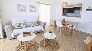 Uma sala de estar moderna com sofá, duas mesas redondas, uma mesa de jantar com cadeiras, área de cozinha e uma janela grande com cortinas.