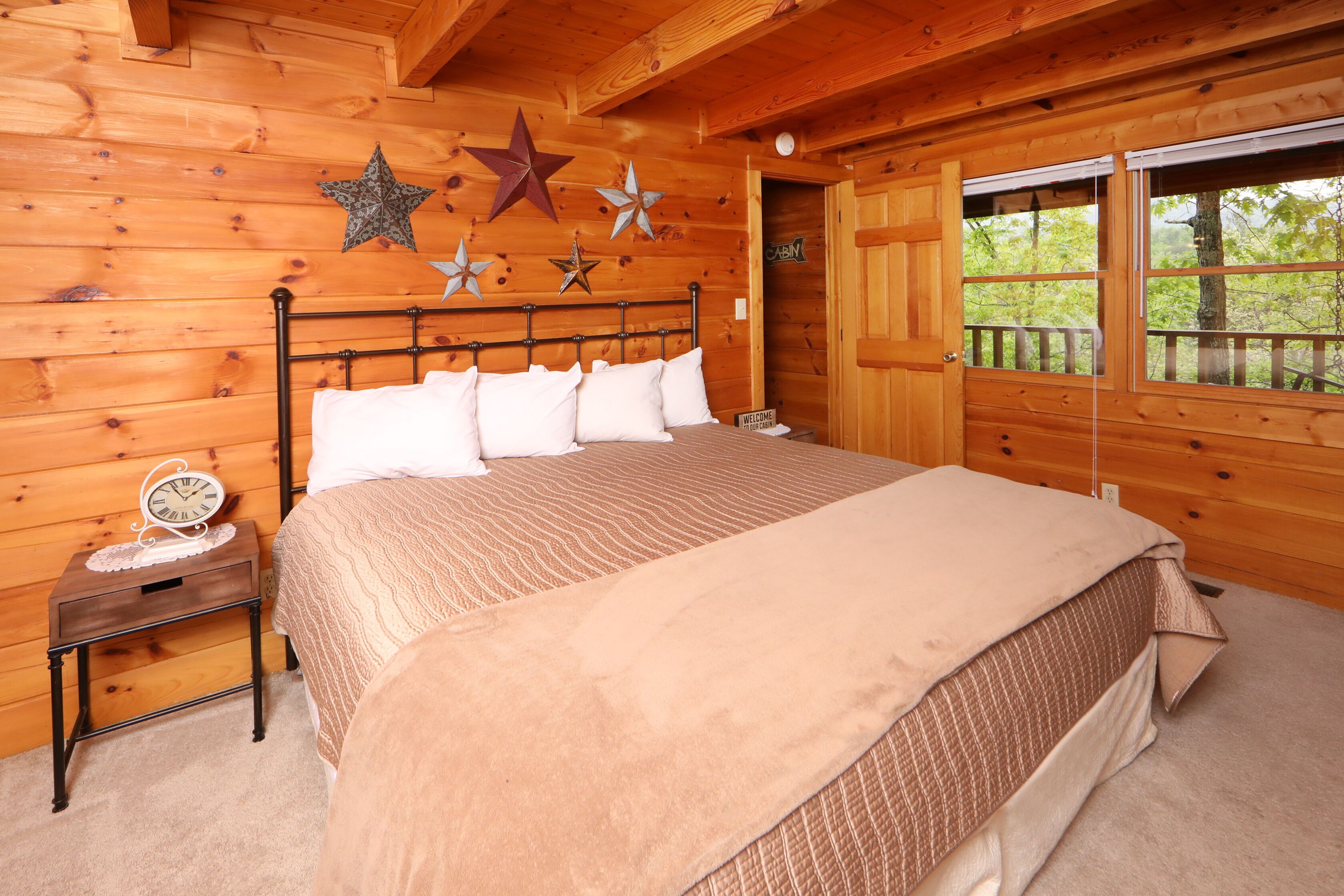 A wooden cabin bedroom with a bed, a nightstand, and a window with a view of trees.