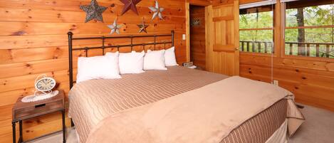 A wooden cabin bedroom with a bed, a nightstand, and a window with a view of trees.