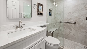 A bathroom with a white sink, mirror, and toilet, a shower with glass doors, and marble walls. - Cozy Garden View 1 bed, 1 bath at Sanibel Moorings #912 (Sanibel)