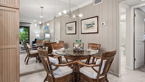 A dining area with a round glass table surrounded by wicker chairs, framed artwork on the walls, and a view into another room. - Cozy Garden View 1 bed, 1 bath at Sanibel Moorings #912 (Sanibel)