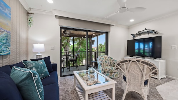 A living room with a sofa, a coffee table, a TV, and a balcony with outdoor furniture. - Cozy Garden View 1 bed, 1 bath at Sanibel Moorings #912 (Sanibel)