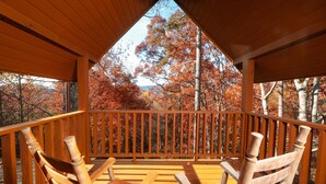 Un patio in legno con due sedie e un tavolo, affacciato su una foresta con alberi dai colori autunnali.