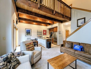 A spacious living area with a wooden ceiling, a kitchen in the background, and a staircase leading to an upper level.