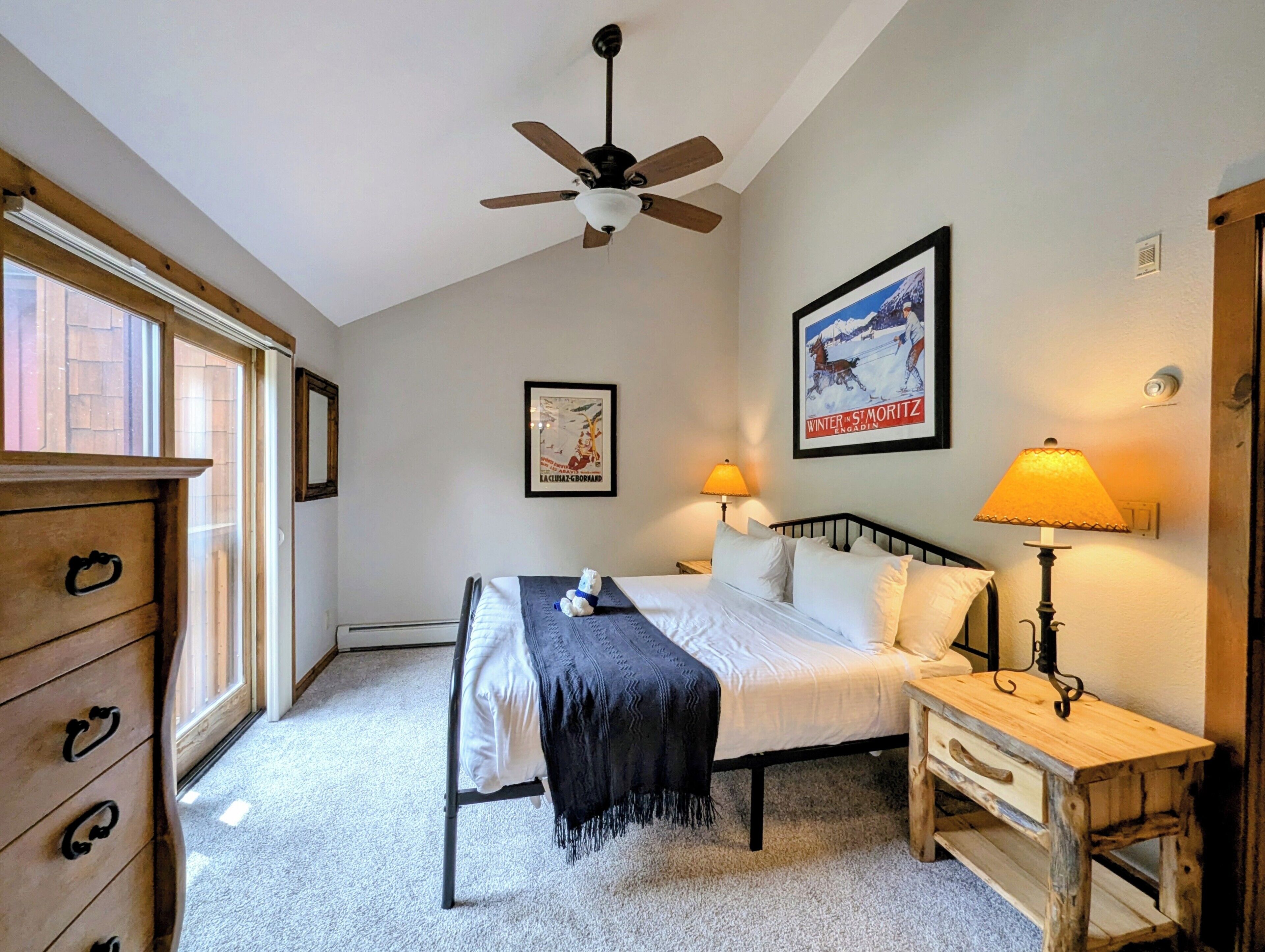 Une chambre à coucher comprenant un lit, une commode en bois, une table de chevet avec une lampe, un ventilateur de plafond et deux tableaux encadrés accrochés au mur.