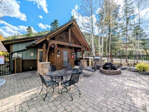A lodge with a patio area featuring a stone fireplace, outdoor seating, and a view of the surrounding forest.