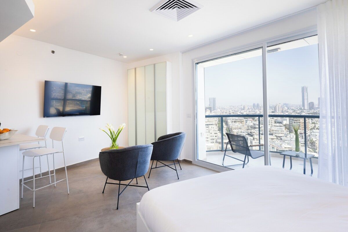 A modern hotel room with a large bed, a desk with chairs, a TV, and a balcony with a city view.