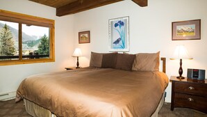 A bedroom with a large bed, two bedside lamps, a framed picture on the wall, and a window with a mountain view.