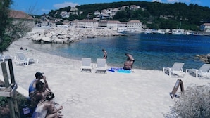 Zona de playa con suelo de piedra, sillones blancos y vista al mar y edificios al fondo.
