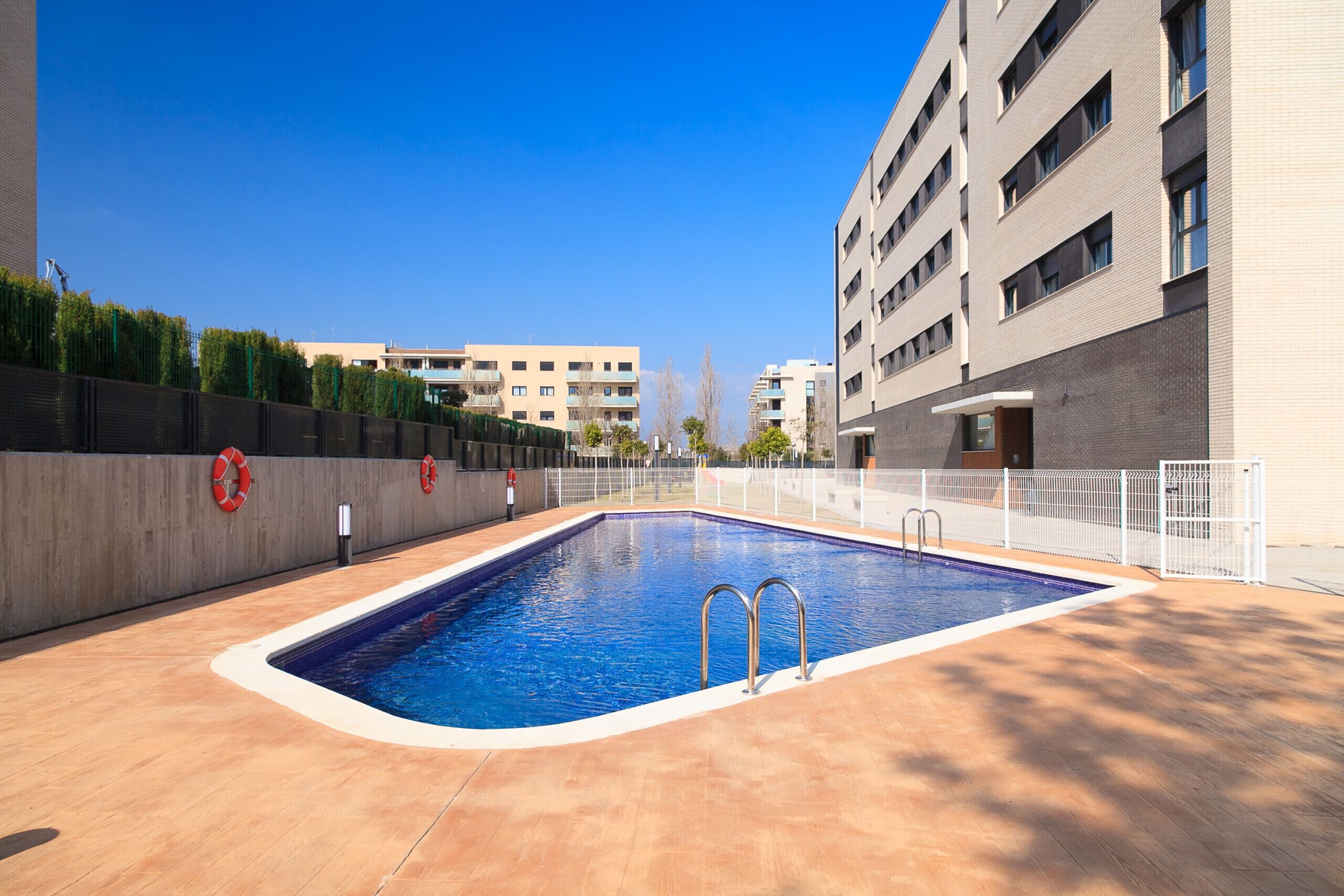 A swimming pool with a tiled deck, surrounded by a white fence, and adjacent to a multi-story building with balconies.