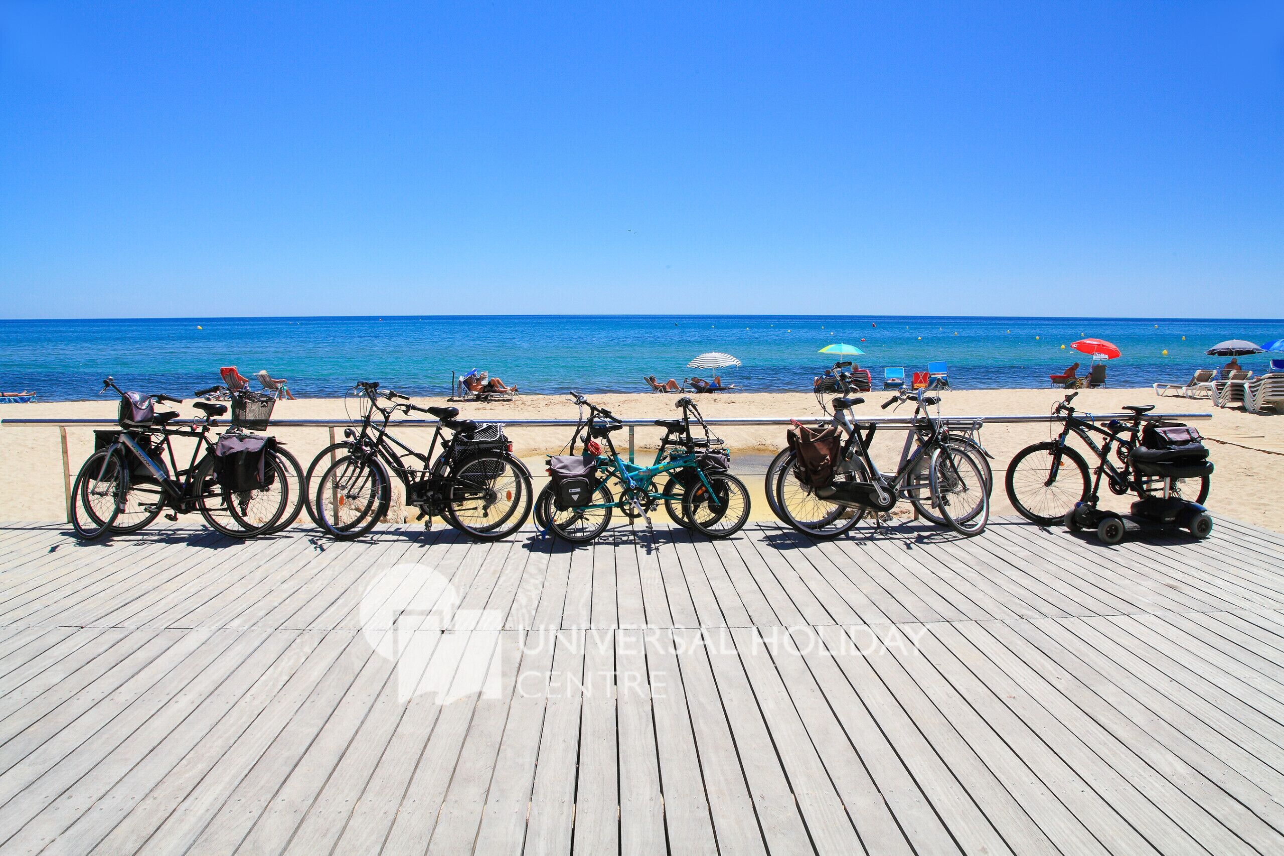 A wooden pier with bicycles parked along it, overlooking a beach with umbrellas and people relaxing.