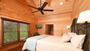 Quarto de cabana de madeira com cama, ventilador de teto, televisão e janela com persianas.