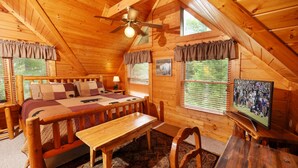 A cozy log cabin bedroom with a bed, bench, TV, and wooden ceiling fan. - New Hot Tub, Beautiful Cabin, Resort Pool, Putt Putt, Golf, 5mi to Pigeon Forge (Sevierville)