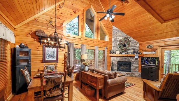 A cozy cabin interior with a wooden dining table, leather sofa, and a stone fireplace. - New Hot Tub, Beautiful Cabin, Resort Pool, Putt Putt, Golf, 5mi to Pigeon Forge (Sevierville)