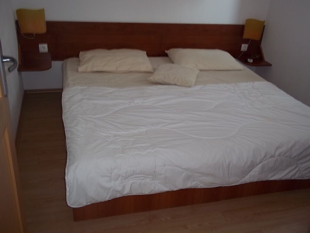 Een net opgemaakt bed met witte lakens en twee kussens, een houten hoofdbord, en nachtlampjes aan beide zijden.