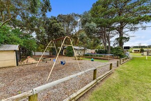 Un parque infantil con columpios, un cobertizo pequeño y una mesa de picnic en una zona verde con árboles.