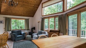 Una habitación acogedora con techo de madera, ventanas grandes con cortinas, un sofá, sillas, una mesita y una alfombra.