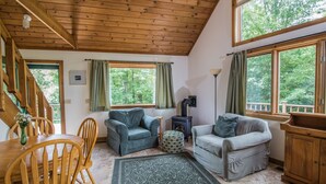 Un cuarto acogedor con vigas de madera en el techo, chimenea y ventanas amplias que dan a un bosque.