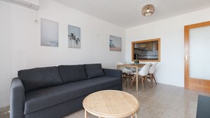 A modern living room with a grey sofa, a wicker table, and a dining area with a wooden table and chairs. - Beachfront apartment perfect for families · UHC ARCO DEL SOL 277 (Hospitalet de L´Infant)