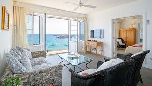 A spacious room with a large window offering a view of the sea, a ceiling fan, a glass coffee table, wicker furniture, and a small desk with a TV. - Casa Lola, Sea Front Apartment in Playa Blanca (Playa Blanca)