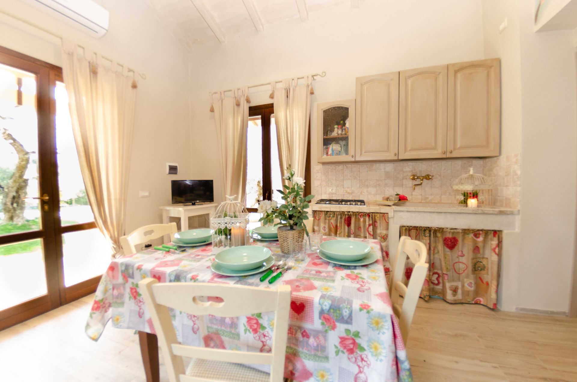 Una cocina acogedora con una mesa de comedor preparada para cuatro personas, un televisor y una ventana con cortinas.