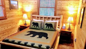 Un dormitorio acogedor en una cabaña de madera con una cama que tiene una colcha con motivos de oso, dos lámparas de noche y un cuadro en la pared.