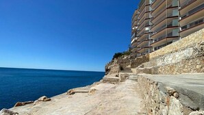 A coastal road with a stone pathway leading to a modern building with balconies overlooking the sea.