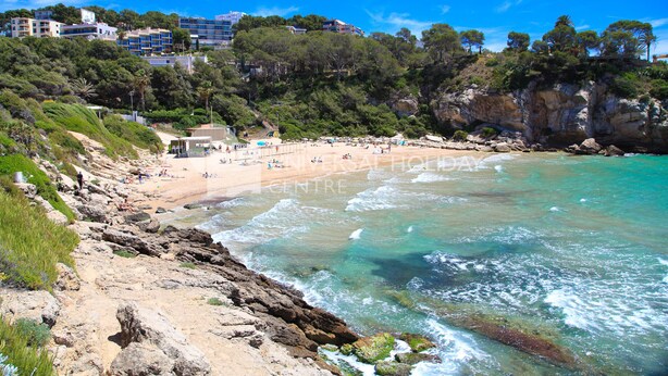 A coastal beach with rocky terrain, clear turquoise waters, and a backdrop of buildings and greenery.