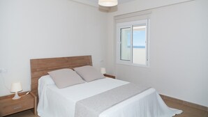 A bedroom with a wooden headboard, a bed with white bedding, a bedside lamp, and a window with a view of the beach.