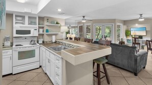 A modern kitchen with white cabinets, a built-in microwave, and a stove. There is a dining area with a gray sofa and a TV on the wall. - Ground Floor, Screened Porch, Free Beach Chair Service, Large Pool (Miramar Beach)