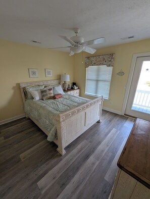 Quarto com cama, ventoinha no teto, mesa de cabeceira, abajur, janela com persianas e porta.