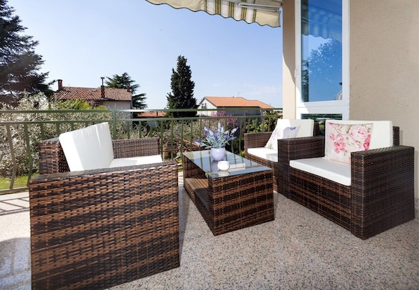 A balcony with wicker furniture, a glass table, and a view of houses and trees.